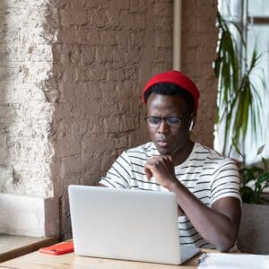 Black freelancer man wear wireless headphones watching webinar on laptop remote online work in cafe